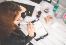 A woman sitting at a desk with a pen in her mouth looking at a notepad