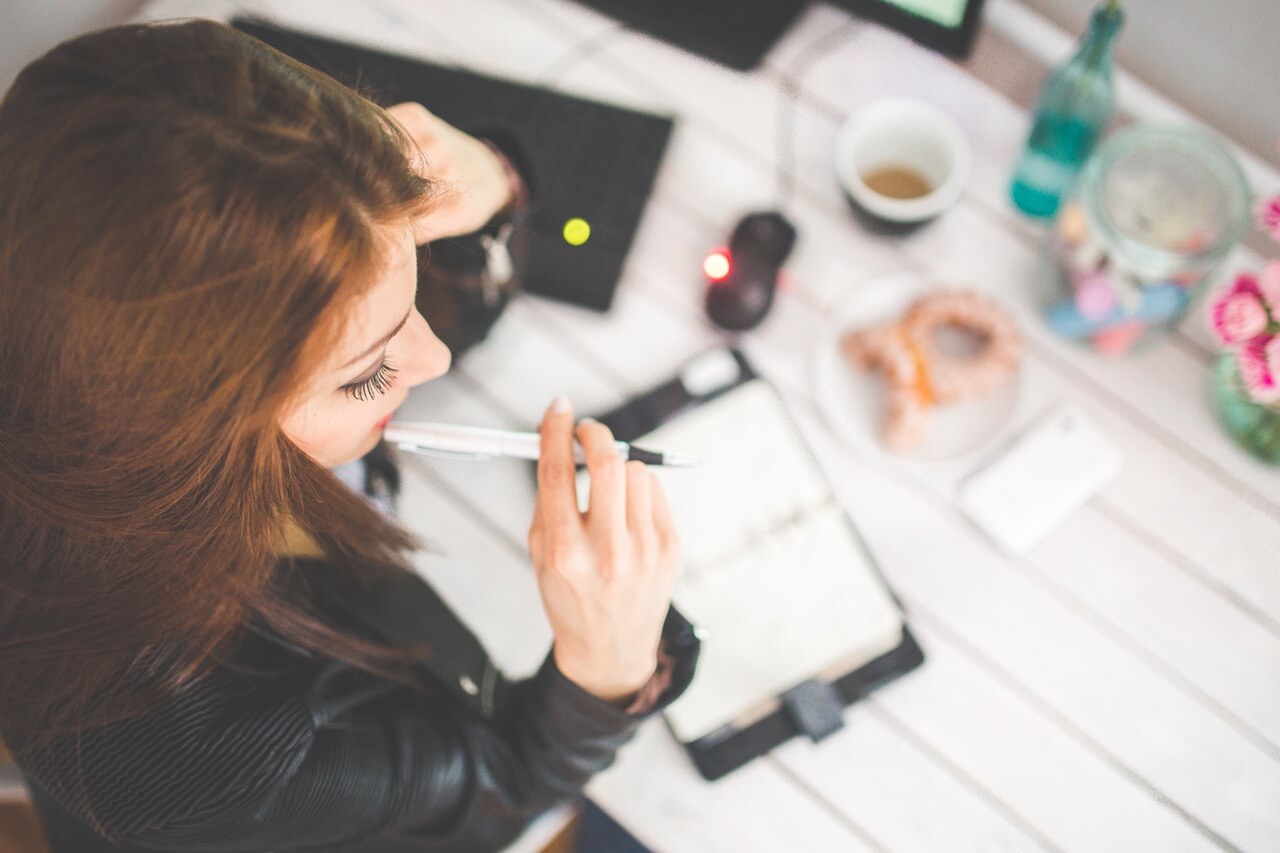 A woman sitting at a desk with a pen in her mouth looking at a notepad
