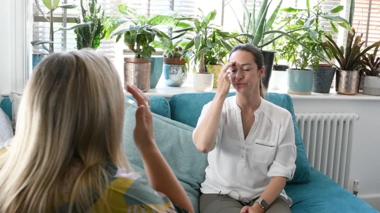 Two women sat on sofa opposite each other tapping their foreheads